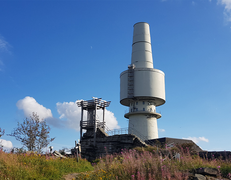 Fernmeldeturm und Aussichtsplattform "Backöfele" auf dem Gipfel des Schneebergs im Fichtelgebirge, umgeben von Felsen, Wiesen und blauer Himmel.